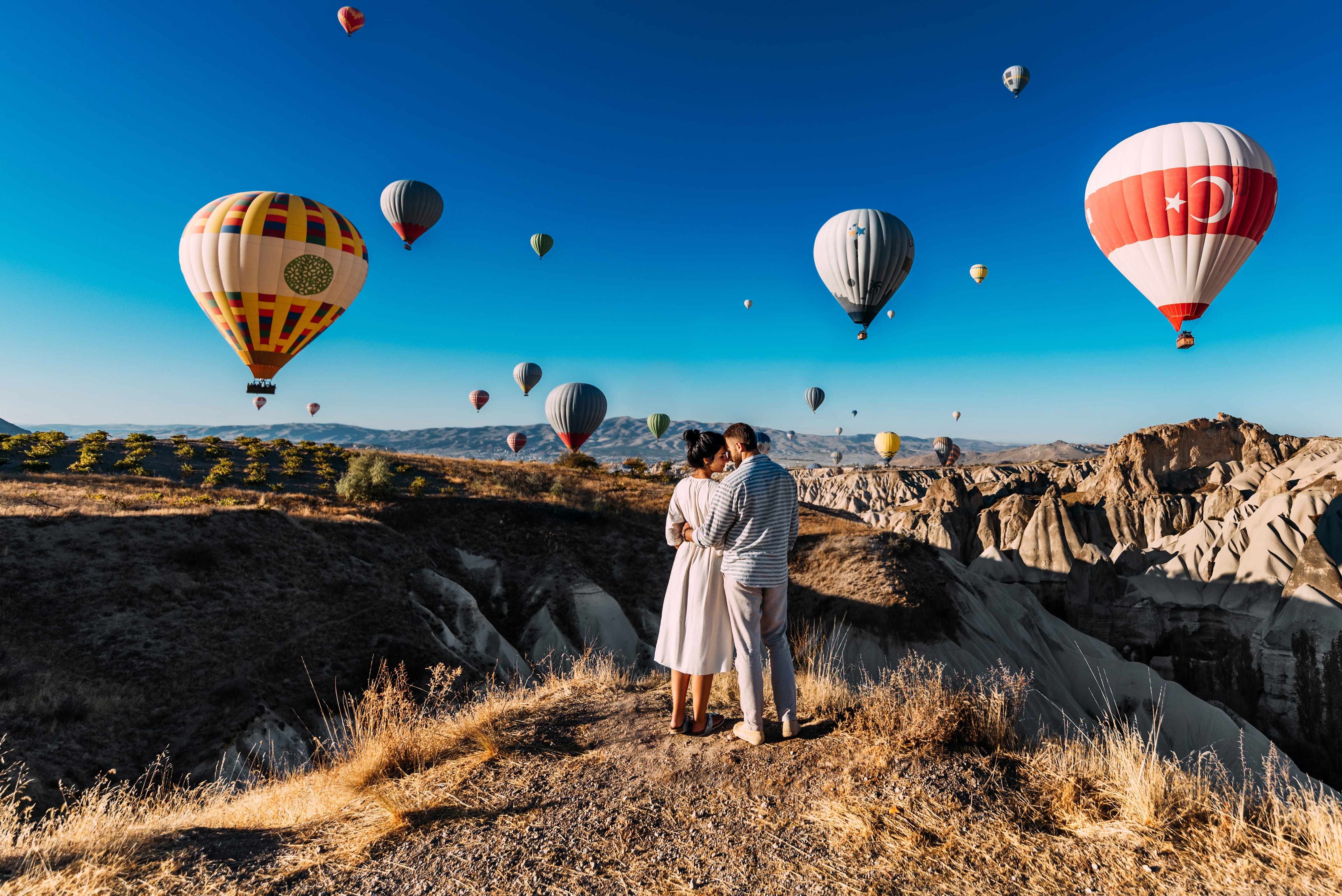 Paar, welches eng umschlungen vor einer schönen Kulisse mit vielen Heißluftballons steht.